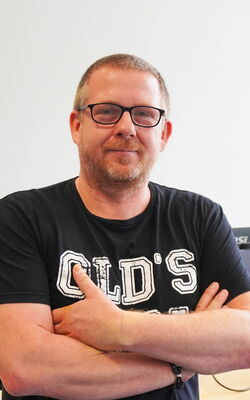 Ein Mann mit Brille und Bart trägt ein schwarzes T-Shirt, während er in einem modernen Büro mit verschränkten Armen lächelt.
