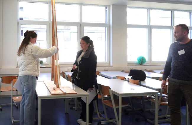 Schülerinnen führen ein Experiment mit einer geneigten Ebene durch, während ein Lehrer sie beobachtet.