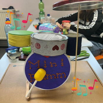 Ein selbstgebautes Mini-Schlagzeug aus Dosen steht auf einem Tisch, während im Hintergrund jemand bastelt.