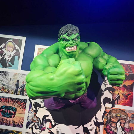 Eine große grüne Figur durchbricht eine Wand aus Comicseiten und wirkt dabei kraftvoll und dynamisch.