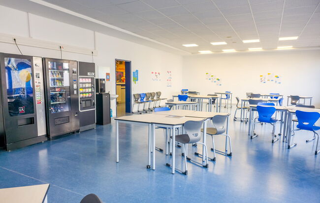 Ein heller Aufenthaltsraum mit Tischen, Stühlen und Verkaufsautomaten bietet Platz für Schüler und kreative Aktivitäten.