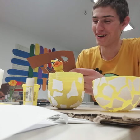 Ein junger Mann in einem gelben T-Shirt bastelt fröhlich an einem gelben Osterküken aus Papiermache.