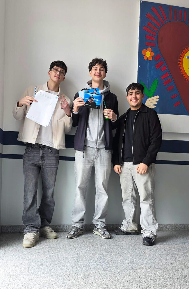 Drei Schüler stehen in einem Schulflur und halten stolz ihre Unterlagen und ein Heft, lächelnd vor einer bunten Wand.