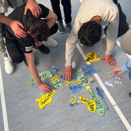 Mehrere Personen arbeiten gemeinsam an einem Bodenpuzzle, das aus bunten, beschrifteten Teilen besteht.