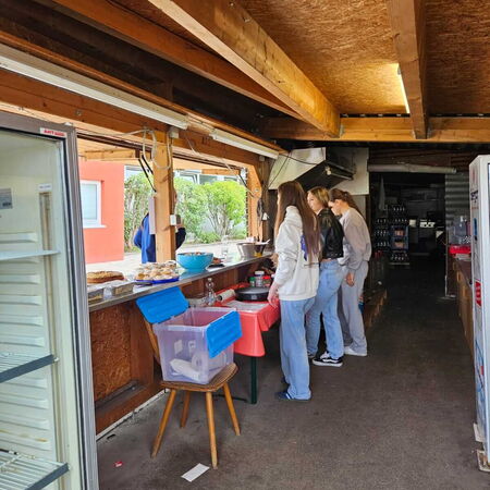 Drei Personen stehen an einem improvisierten Verkaufsstand mit Kuchen und Getränken in einem offenen Holzpavillon.