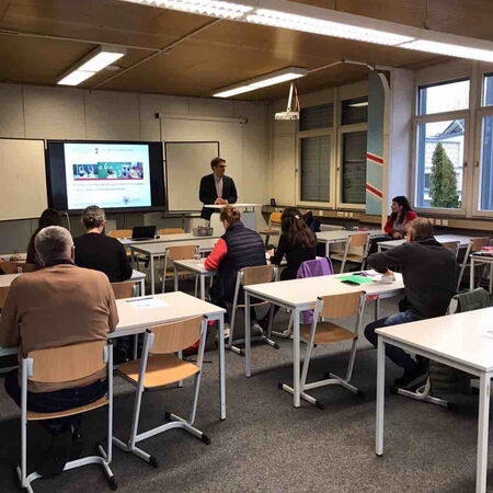 In einem Klassenzimmer hält ein Lehrer eine Präsentation vor einer Gruppe aufmerksam lauschender Erwachsener.