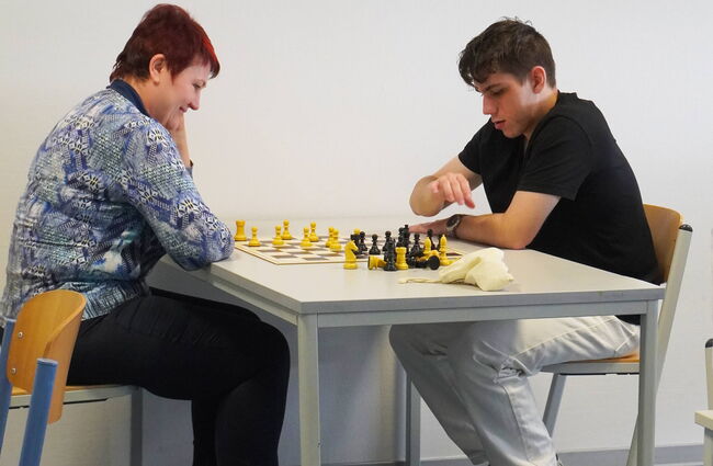 Eine Lehrkraft und ein Schüler spielen konzentriert Schach an einem Tisch in einem Klassenzimmer.