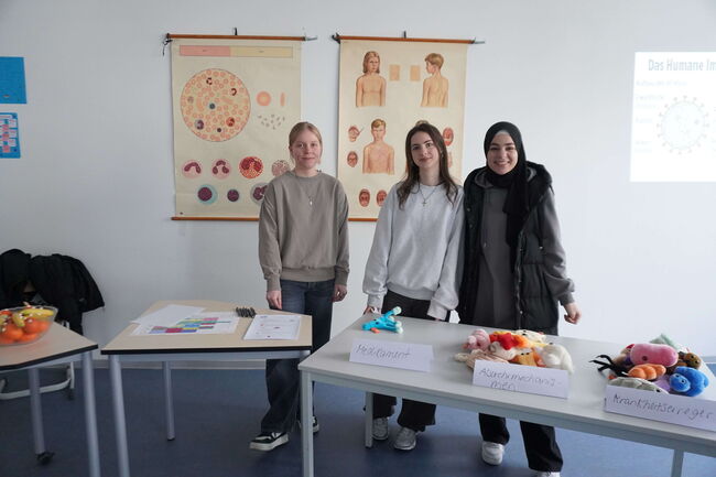 Drei Personen stehen in einem Klassenzimmer vor Tischen mit Lernmaterialien und Postern zur menschlichen Anatomie.