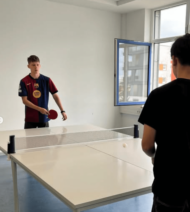 Zwei Jugendliche spielen Tischtennis in einem hellen Raum, während ein Fenster geöffnet ist und Licht hereinfällt.