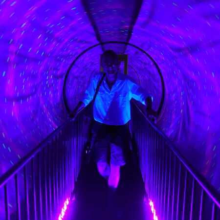 Eine Person geht durch einen leuchtenden, violetten Tunnel mit bunten Lichtpunkten, die eine futuristische Atmosphäre schaffen.