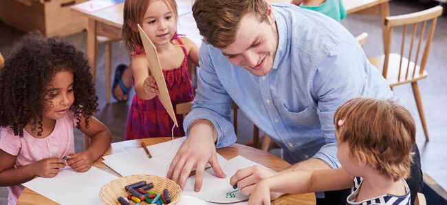 Ein Erzieher sitzt mit drei Kita-Kindern am Tisch