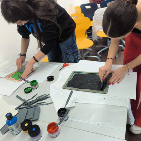 Zwei Schülerinnen arbeiten an einem Tisch und drucken mit Farben auf ein T-Shirt in einem kreativen Workshop.