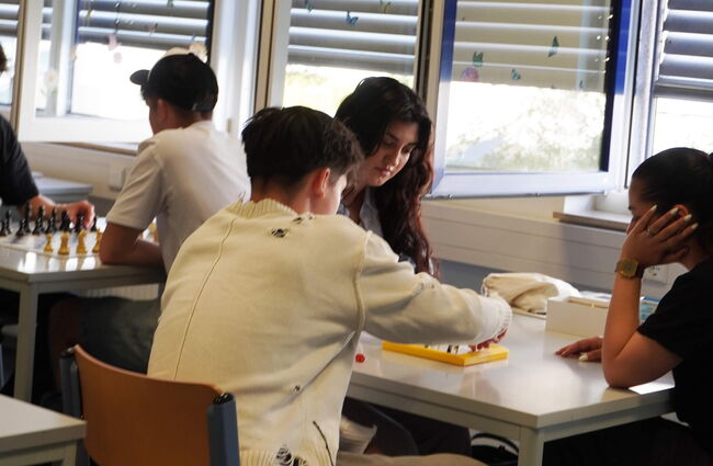 Schüler spielen Schach und ein Brettspiel in einem hellen Klassenzimmer mit großen Fenstern.