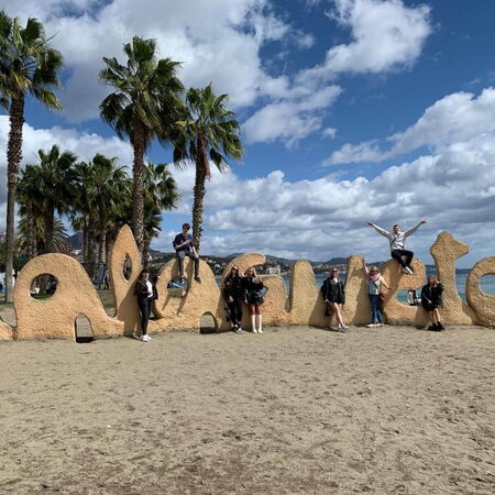 Menschen posieren fröhlich auf dem großen "Malagueta"-Schriftzug am Strand von Málaga unter Palmen und blauem Himmel.