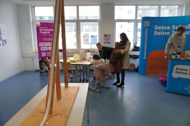 Ein Klassenzimmer mit Informationsständen, an denen sich Erwachsene und Kinder umsehen und unterhalten.