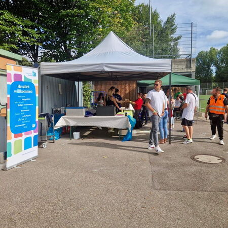 Menschen versammeln sich unter einem Pavillon bei einem Willkommenstreffen neben einem Fußballplatz.