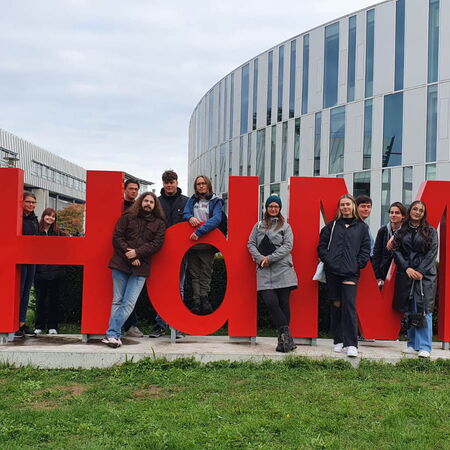 Eine Gruppe von Schülern posiert fröhlich vor einem großen roten "HdM"-Schriftzug auf einer Wiese.