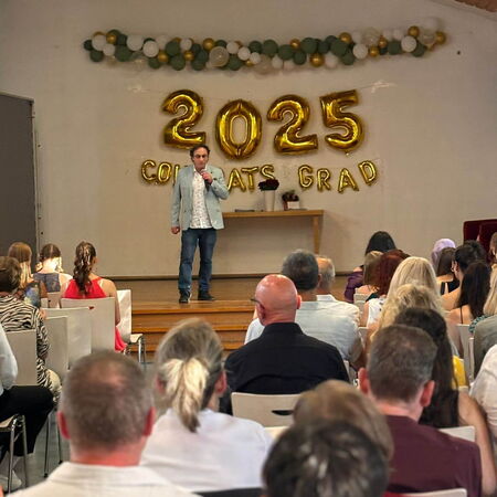Ein Redner steht auf einer Bühne vor einer "2025 Congrats Grad"-Dekoration, während das Publikum aufmerksam zuhört.