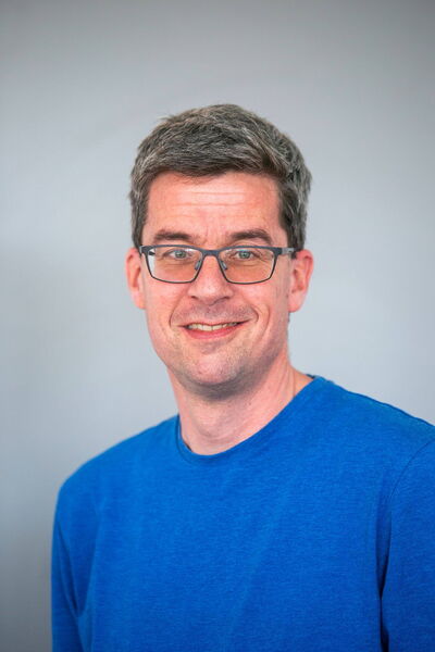 Ein Mann in einem blauen T-Shirt lächelt freundlich in die Kamera vor einem neutralen Hintergrund.