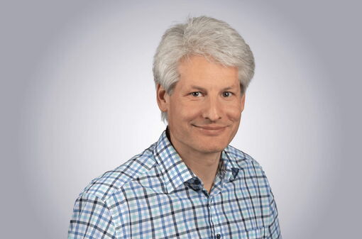 Ein lächelnder Mann in kariertem Hemd vor einem neutralen Hintergrund.