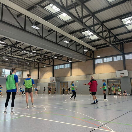 In einer Sporthalle spielen Jugendliche ein intensives Handballspiel unter der Leitung eines Trainers in rotem Trikot.