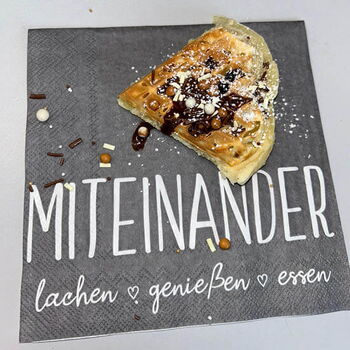 Ein mit Schokoladensauce und Streuseln belegtes Kipferl liegt auf einer Serviette mit der Aufschrift "Miteinander lachen genießen essen".