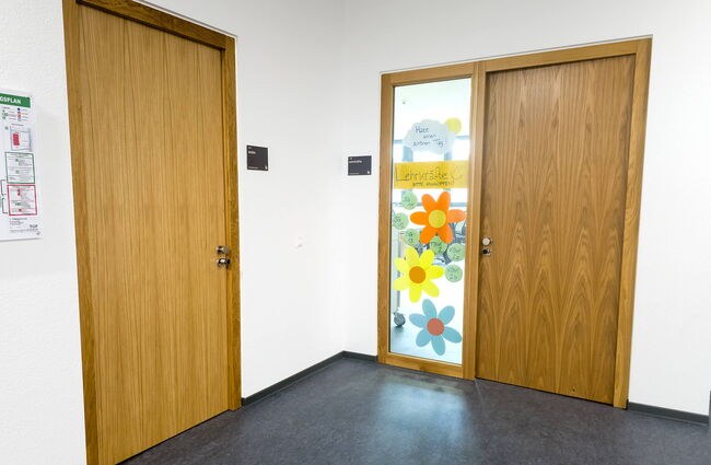 Zwei Holztüren führen in einen Flur, einer mit bunten Blumen und einem Schild, das Lehrer willkommen heißt.