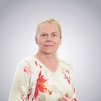 Eine lächelnde Frau in einem geblümten Oberteil steht vor einem neutralen Hintergrund.