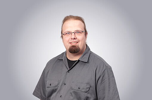 Ein Mann mit Bart und Brille trägt ein graues Hemd und lächelt vor einem neutralen Hintergrund.