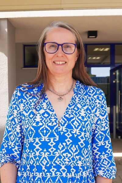 Eine lächelnde Frau mit langen Haaren und blauer Bluse steht vor einem Gebäude mit Glasdoor.