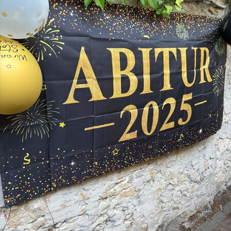 Ein Banner mit der Aufschrift "Abitur 2025" hängt an einer Wand, dekoriert mit bunten Luftballons und Feuerwerksmotiven.