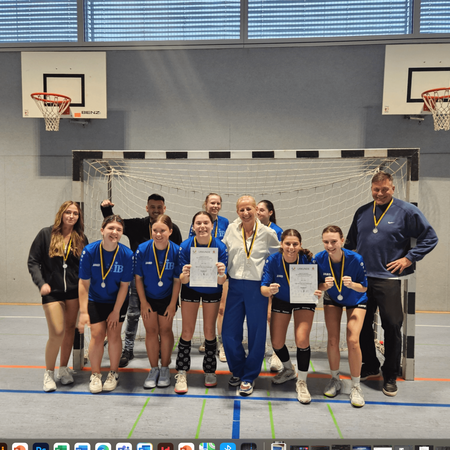 Eine Gruppe von jungen Athletinnen mit Medaillen posiert in einer Sporthalle vor einem Handballtor für ein Siegerfoto.