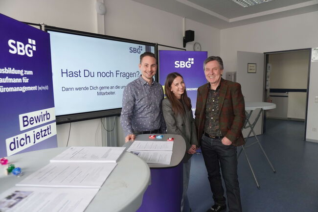 Drei Personen stehen lächelnd vor einem Informationsstand von SBG mit dem Slogan "Bewirb dich jetzt!" im Hintergrund.