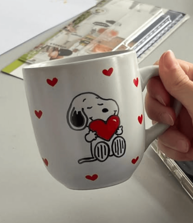 Eine Hand hält eine Tasse mit Snoopy, der ein Herz umgeben von roten Herzen umarmt.