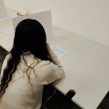 Ein Mädchen mit langen Haaren sitzt an einem Tisch, konzentriert auf ein Blatt Papier vor sich.
