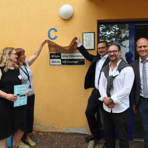Eine Gruppe von Menschen steht vor einer Schule mit einem „Schule ohne Rassismus“ Schild und enthüllt es feierlich.