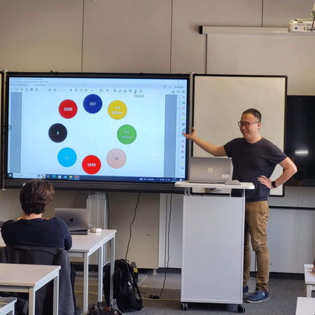 Ein Mann hält eine Präsentation vor einer Klasse und zeigt bunte Zahlen auf einem großen Display.