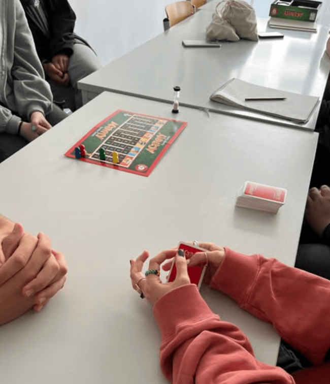 Eine Gruppe von Jugendlichen spielt ein Brettspiel, während Karten und eine Sanduhr auf dem Tisch liegen.
