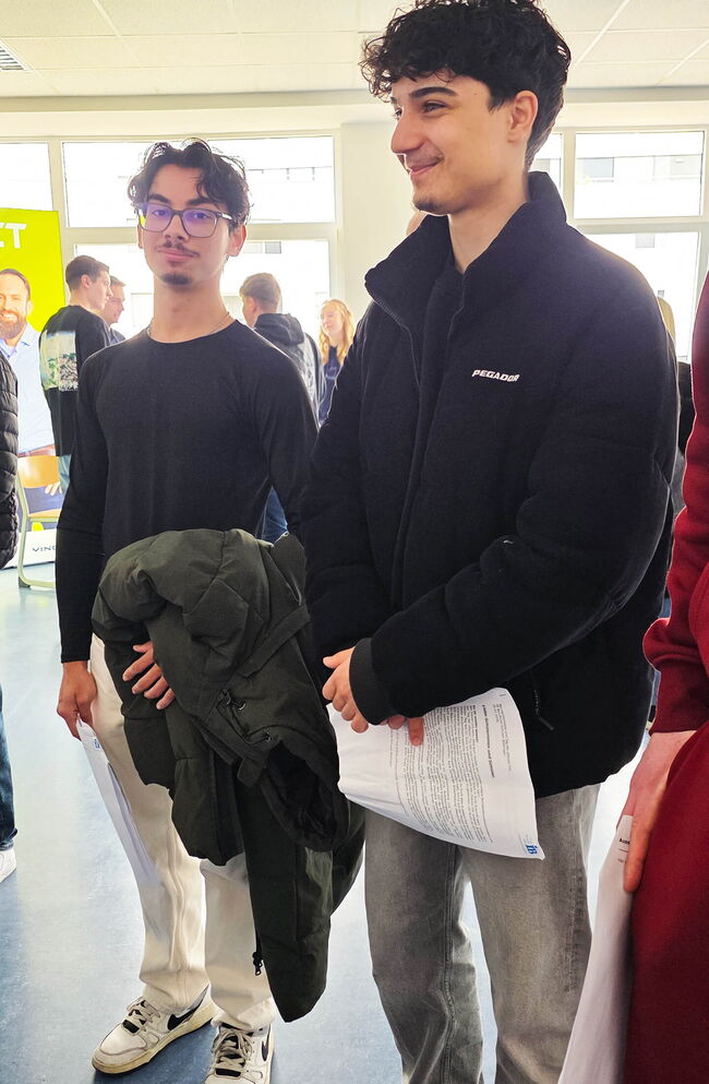 Zwei Schüler stehen in einer Halle, einer hält eine Jacke und Unterlagen, während sie miteinander sprechen.