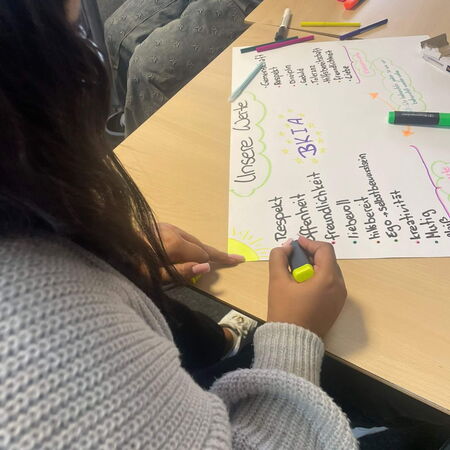 Eine Person schreibt mit einem gelben Stift auf ein Plakat mit dem Titel "Unsere Werte" in einem Klassenzimmer.