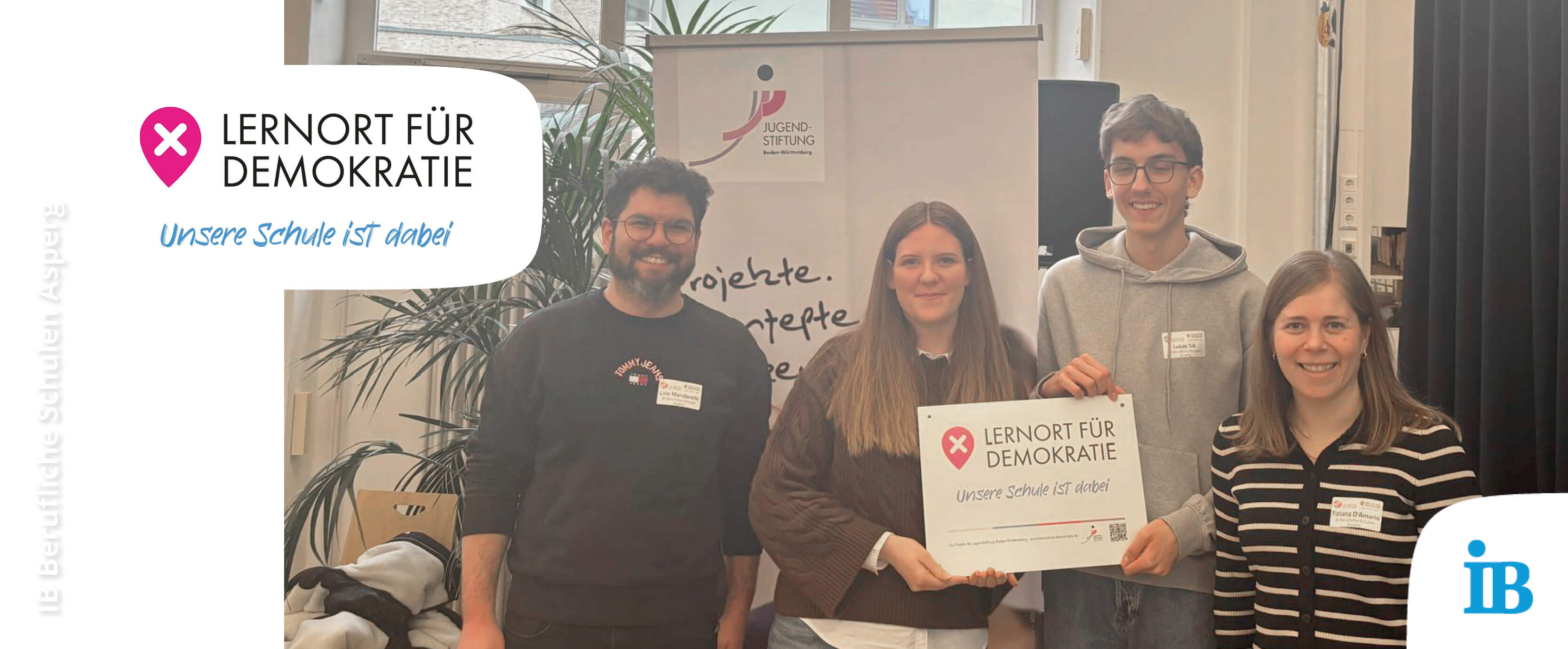 Vier junge Menschen halten ein Schild mit der Aufschrift „Lernort für Demokratie“ in einer freundlichen Umgebung.