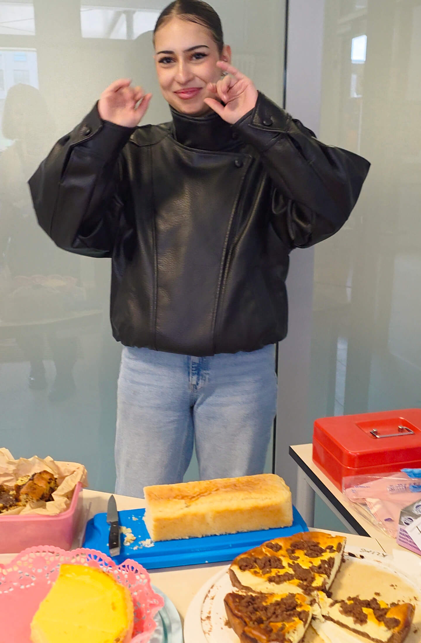 Eine Person in einem schwarzen Lederoberteil lächelt vor einem Tisch mit verschiedenen Kuchen und Backwaren.