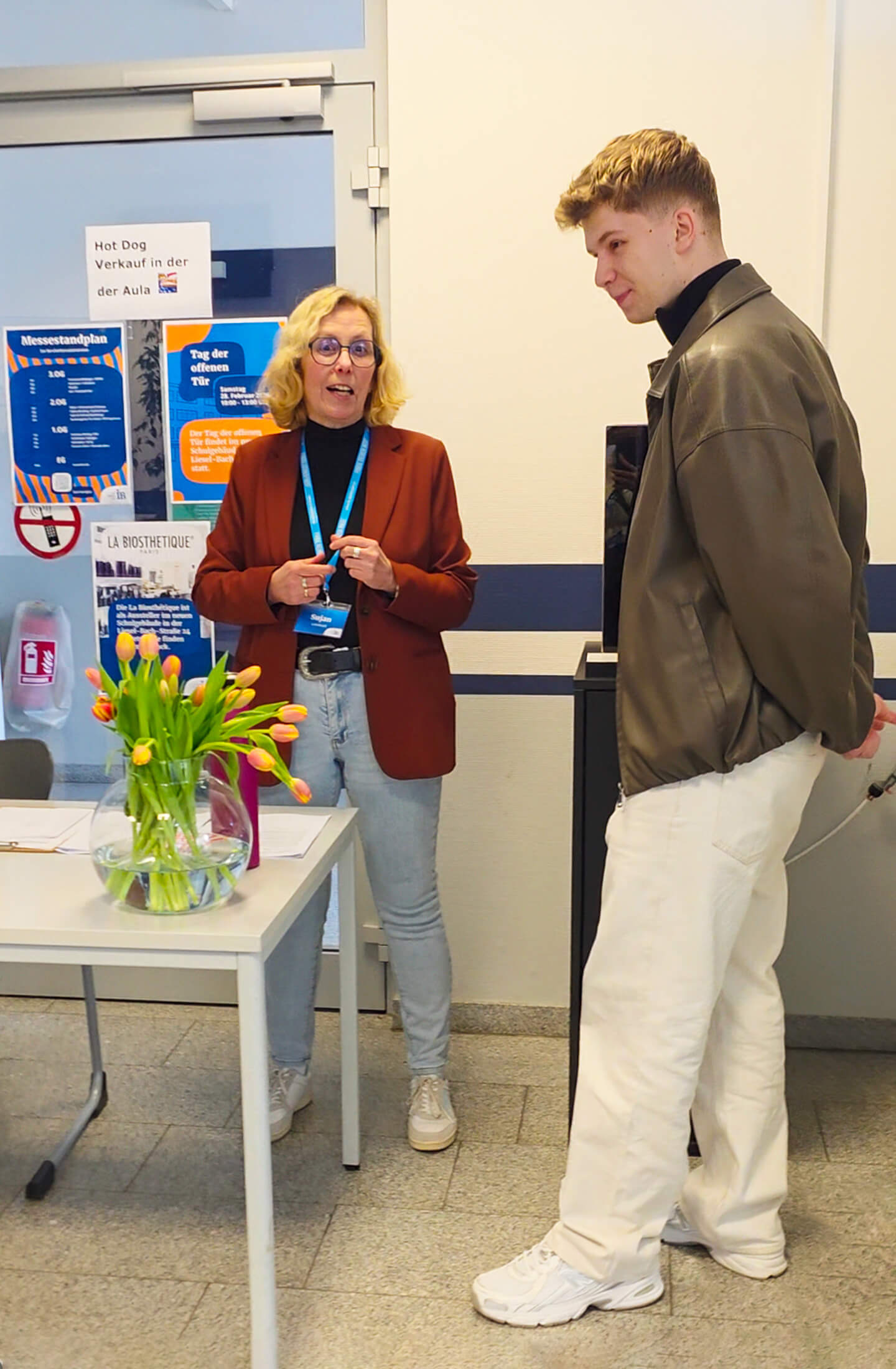 Eine Frau spricht mit einem jungen Mann am Empfang, während ein Blumenstrauß auf dem Tisch steht.