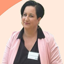 Eine Frau mit kurzem dunklem Haar trägt einen rosa Blazer und ein Namensschild vor einem pastellfarbenen Hintergrund.