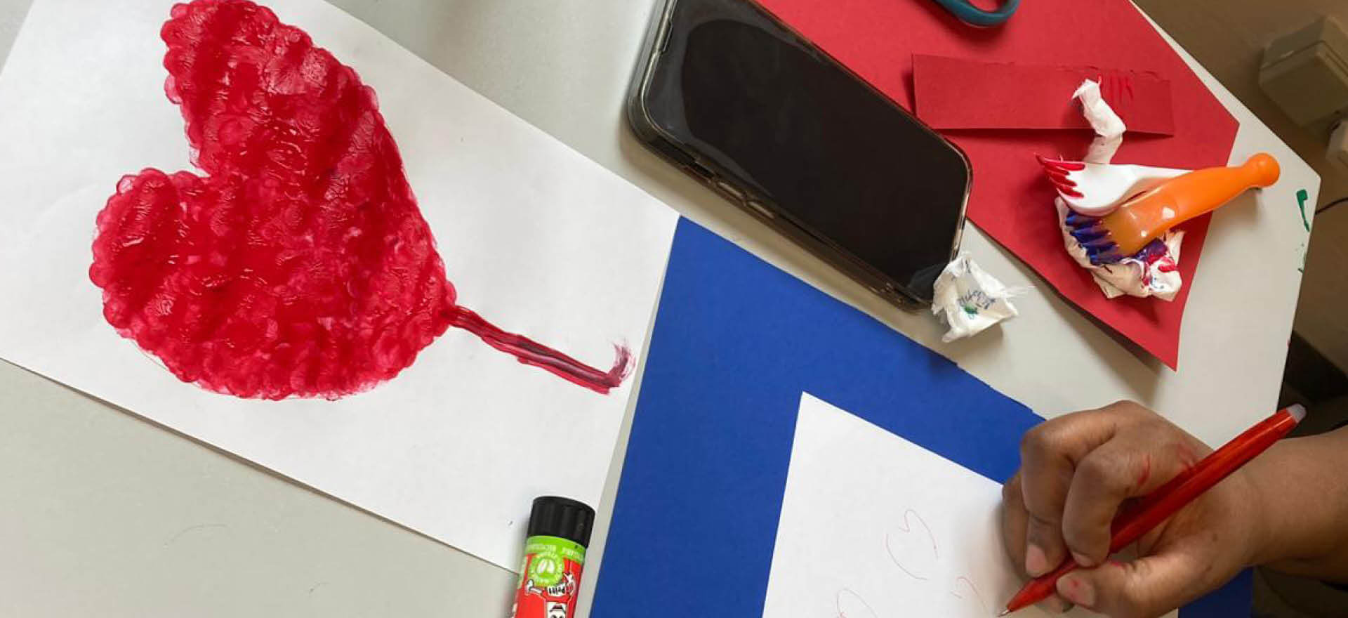 Eine Person zeichnet Herzen auf Papier, umgeben von einem roten Herzbild, Bastelutensilien und einem Handy zum Muttertag.