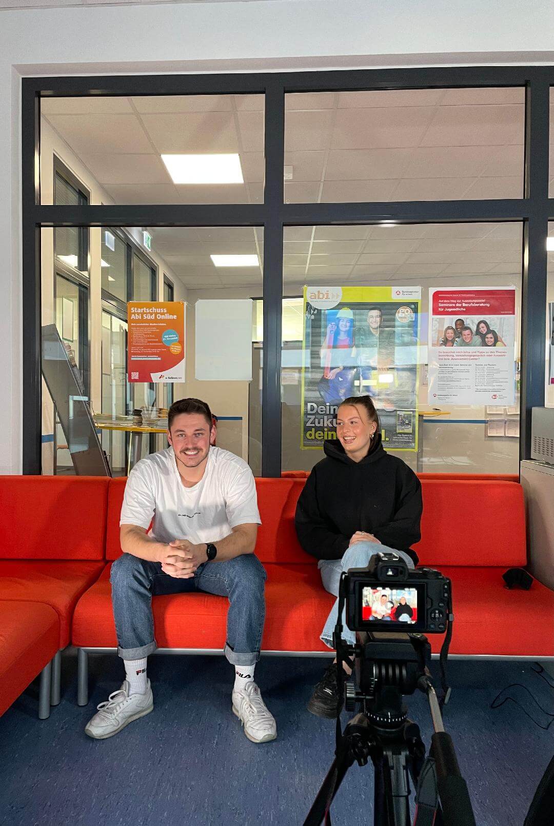 Zwei Personen sitzen auf einem roten Sofa und werden für ein Interview von einer Kamera aufgenommen.