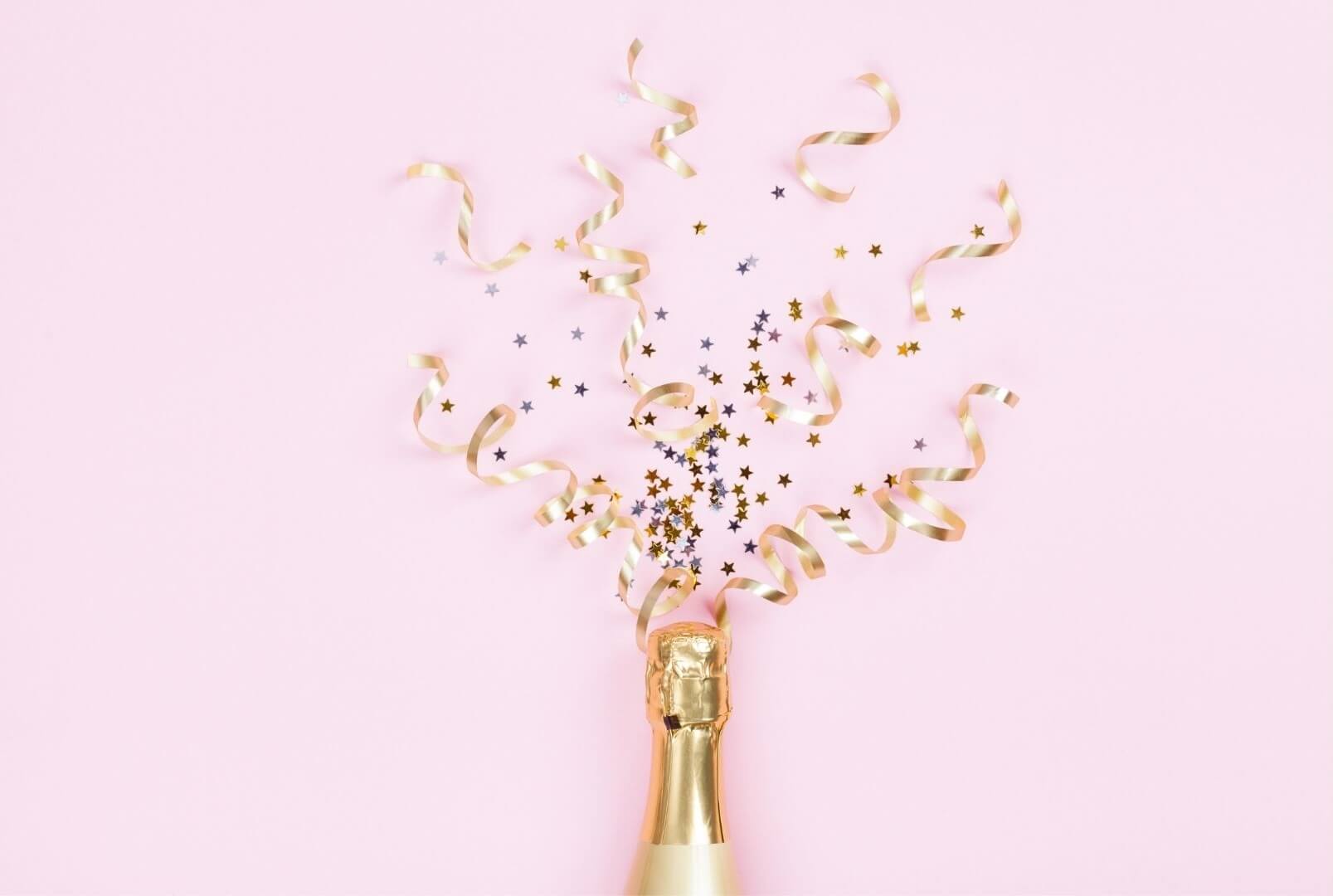 Eine Champagnerflasche auf rosa Hintergrund sprüht goldene Luftschlangen und Sterne zur Feier in die Luft.