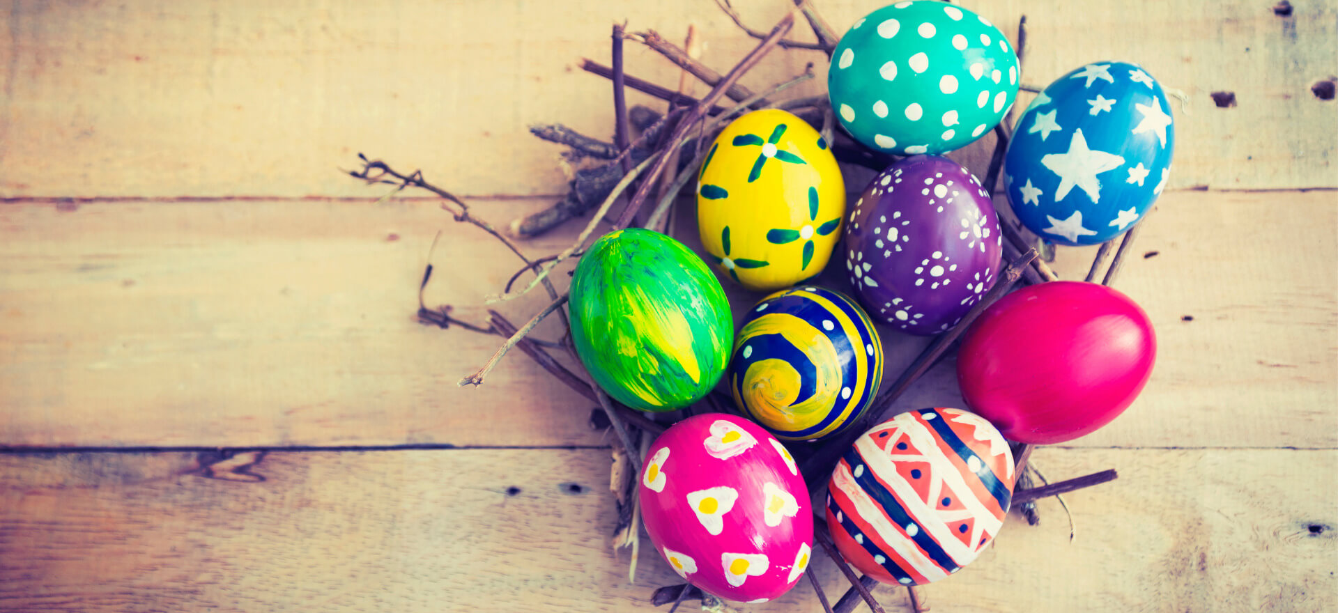 Bunte Ostereier liegen dekorativ in einem Nest aus Zweigen auf einem Holztisch und vermitteln festliche Urlaubsstimmung.