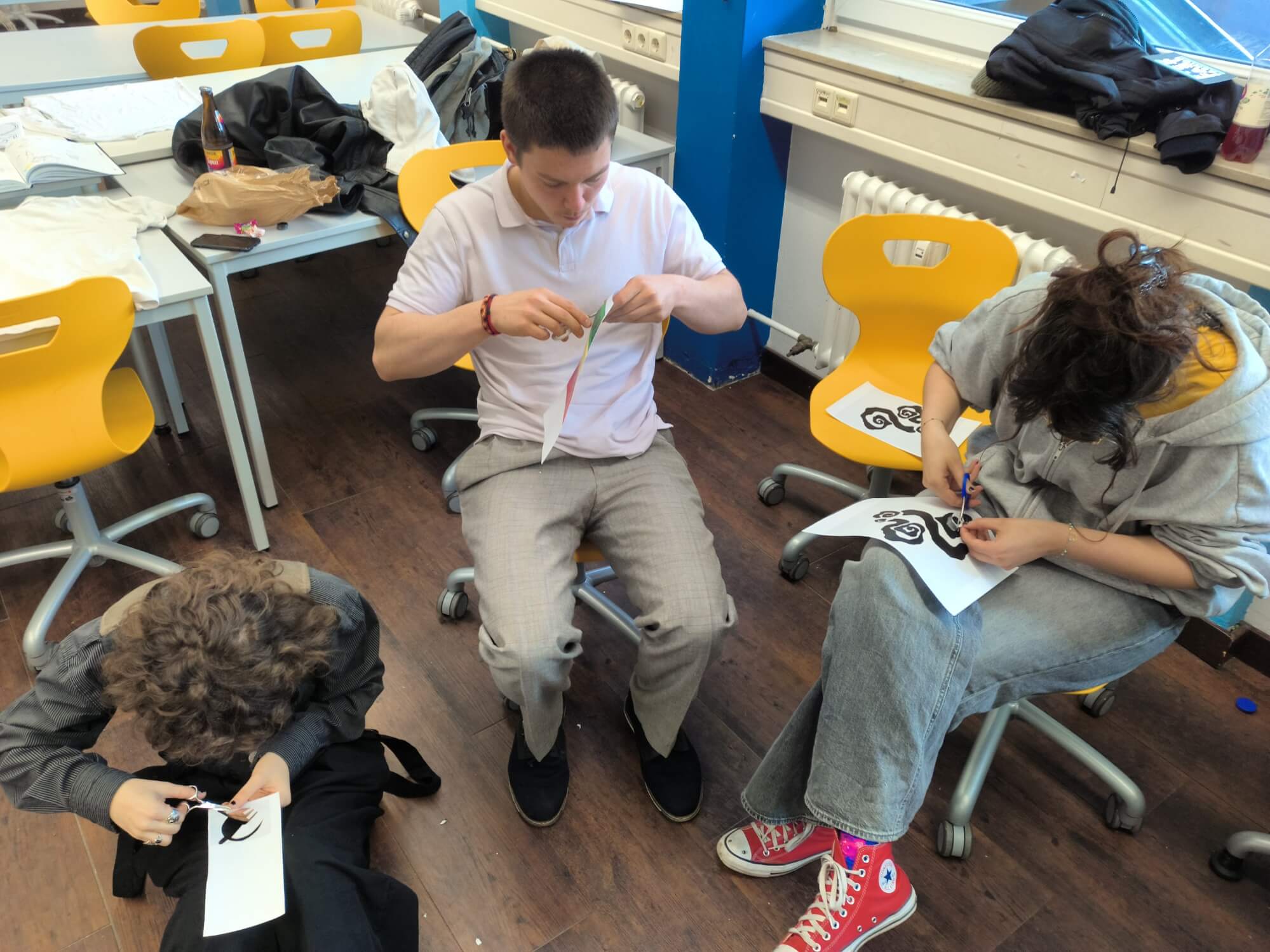 Drei Schüler schneiden konzentriert mit Scheren an Papieren in einem kreativen Klassenraum.
