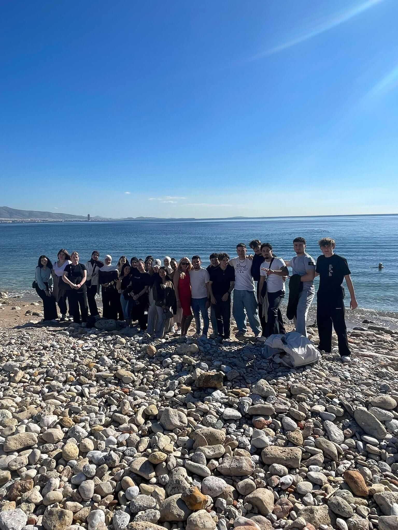 Eine Gruppe von Menschen steht auf einem steinigen Strand vor einem klaren blauen Himmel und Meer am 19. November 2025.